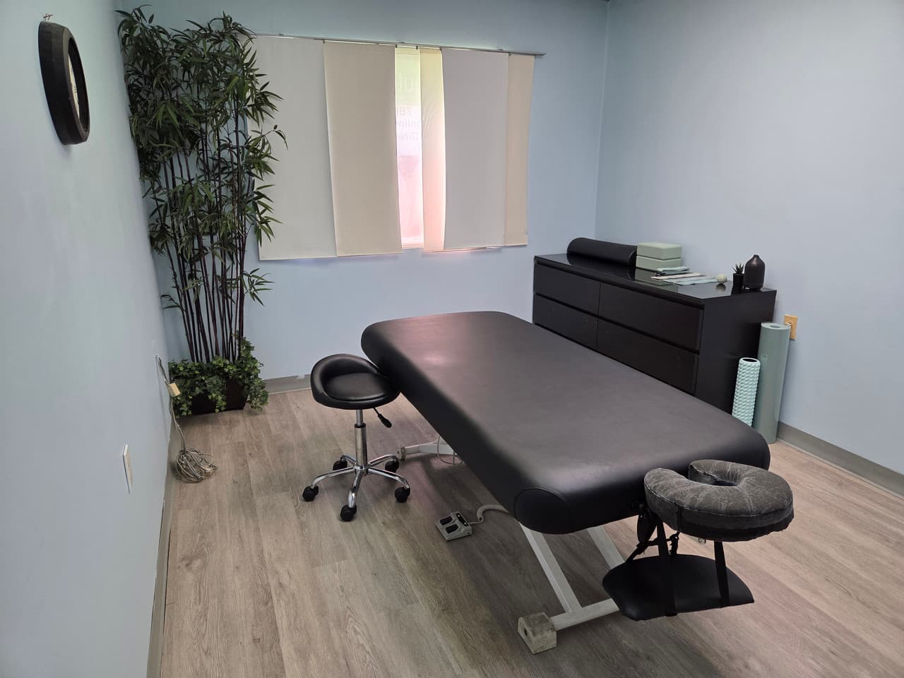 Minimalist massage therapy room featuring a black table, rolling stool, and tall artificial plant.